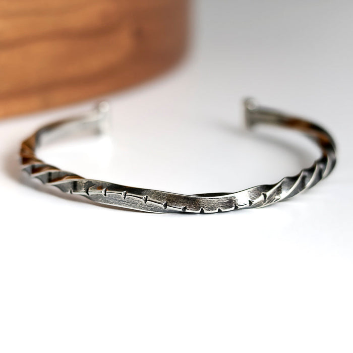 Silver twisted cuff bracelet on a white surface with a wooden object in the background