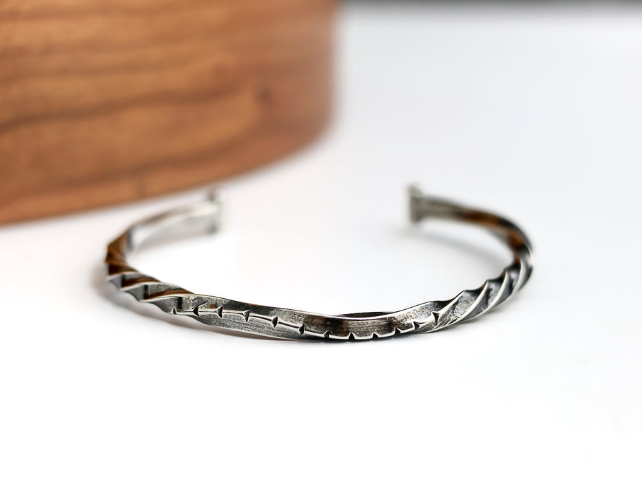 Silver twisted cuff bracelet on a white surface with a wooden object in the background